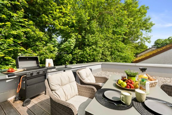 Terrasse mit Grill, gemütlichen Stühlen und gedecktem Tisch mit Obst und Getränken.