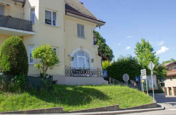 Gelbes Haus an einer Kurve, umgeben von Bäumen und Rasen, blauer Himmel im Hintergrund.