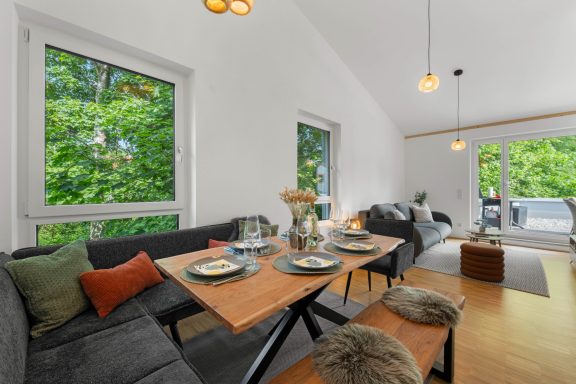 Helles Wohnzimmer mit einem Esstisch, Couch und großen Fenstern, umgeben von Bäumen.